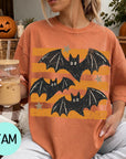 A woman wearing a t-shirt with a bat design is sitting on a couch, holding a drink in a glass. There are pumpkins and a pumpkin decoration in the background, suggesting a Halloween theme.