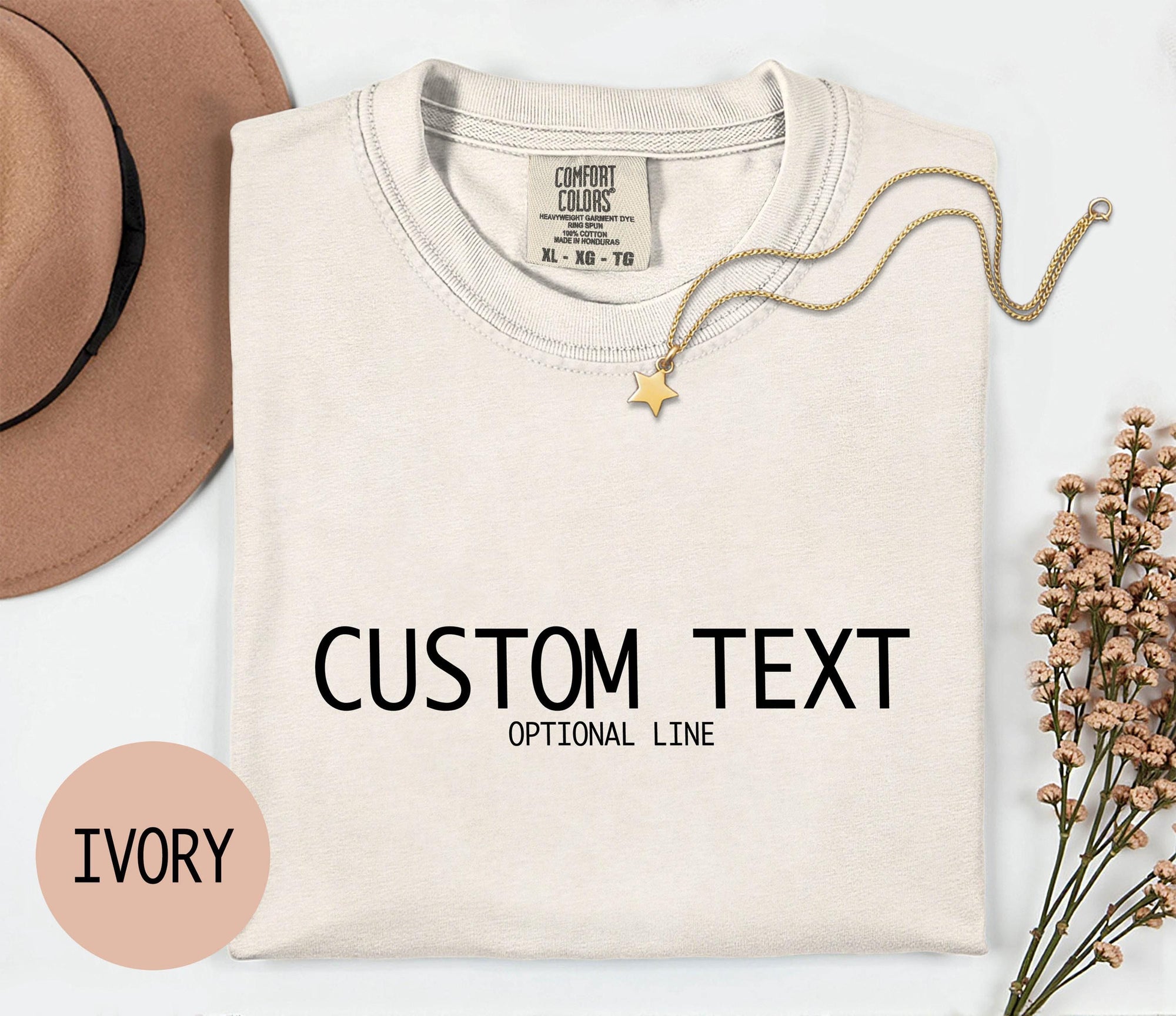 a white t-shirt with the text &amp;quot;CUSTOM TEXT&amp;quot; printed on it, along with the text &amp;quot;OPTIONAL LINE&amp;quot; below it. The t-shirt is displayed on a white background, accompanied by a beige hat, a gold chain, and some pink flowers.