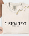 a white t-shirt with the text "CUSTOM TEXT" printed on it, along with the text "OPTIONAL LINE" below it. The t-shirt is displayed on a white background, accompanied by a beige hat, a gold chain, and some pink flowers.