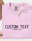 a pink t-shirt with the text "CUSTOM TEXT" and "OPTIONAL LINE" printed on it, along with a necklace and a hat.