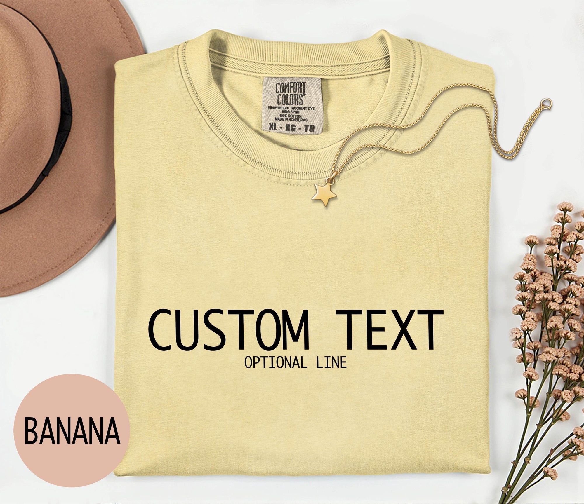 a yellow t-shirt with the text &amp;quot;CUSTOM TEXT&amp;quot; printed on it, along with the word &amp;quot;banana&amp;quot; written below it. The t-shirt is displayed on a white background, and there is a hat and some flowers in the background.