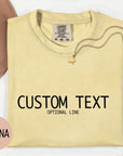 a yellow t-shirt with the text "CUSTOM TEXT" printed on it, along with the word "banana" written below it. The t-shirt is displayed on a white background, and there is a hat and some flowers in the background.