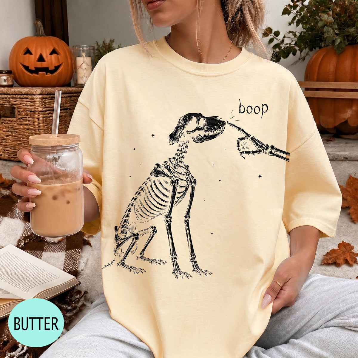A woman wearing a yellow t-shirt with a skeleton graphic and the word "boop" printed on it, holding a drink in a glass.