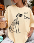 A woman wearing a yellow t-shirt with a skeleton graphic and the word "boop" printed on it, holding a drink in a glass.