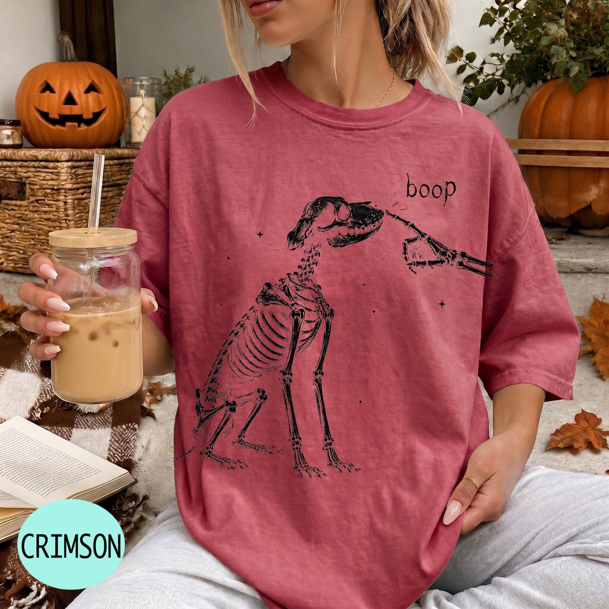 A woman wearing a red t-shirt with a skeleton drawing and the word "boop" printed on it, holding a glass of iced coffee.