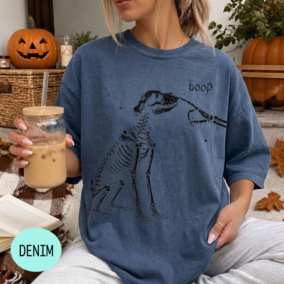A woman wearing a blue t-shirt with a skeleton drawing and the word "boop" printed on it, holding a glass of iced coffee.