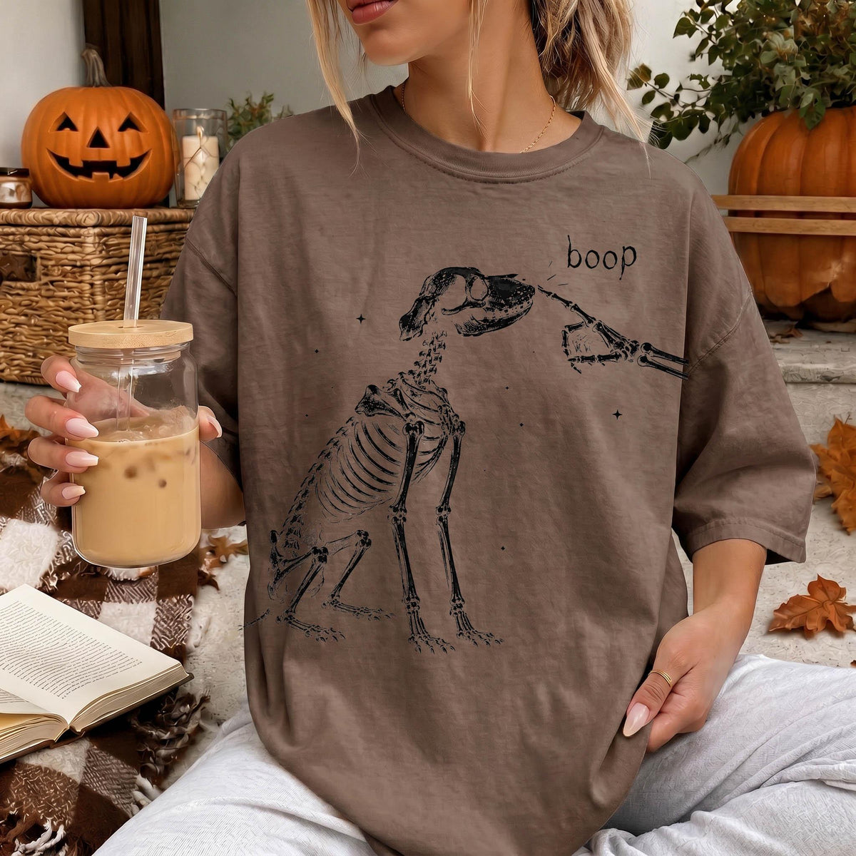 A woman wearing a t-shirt with a skeleton drawing and the word "boop" is sitting on a couch, holding a glass of iced coffee.