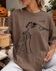 A woman wearing a t-shirt with a skeleton drawing and the word "boop" is sitting on a couch, holding a glass of iced coffee.