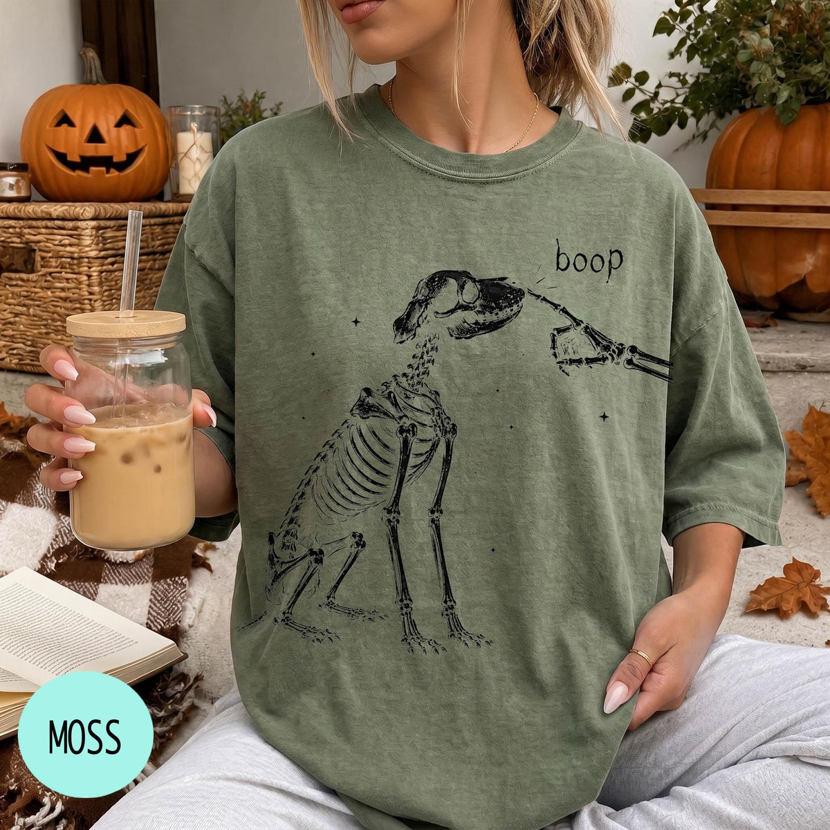 A person wearing a green t-shirt with a skeleton drawing and the word &amp;quot;boop&amp;quot; printed on it, holding a glass of iced coffee.