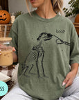 A person wearing a green t-shirt with a skeleton drawing and the word "boop" printed on it, holding a glass of iced coffee.