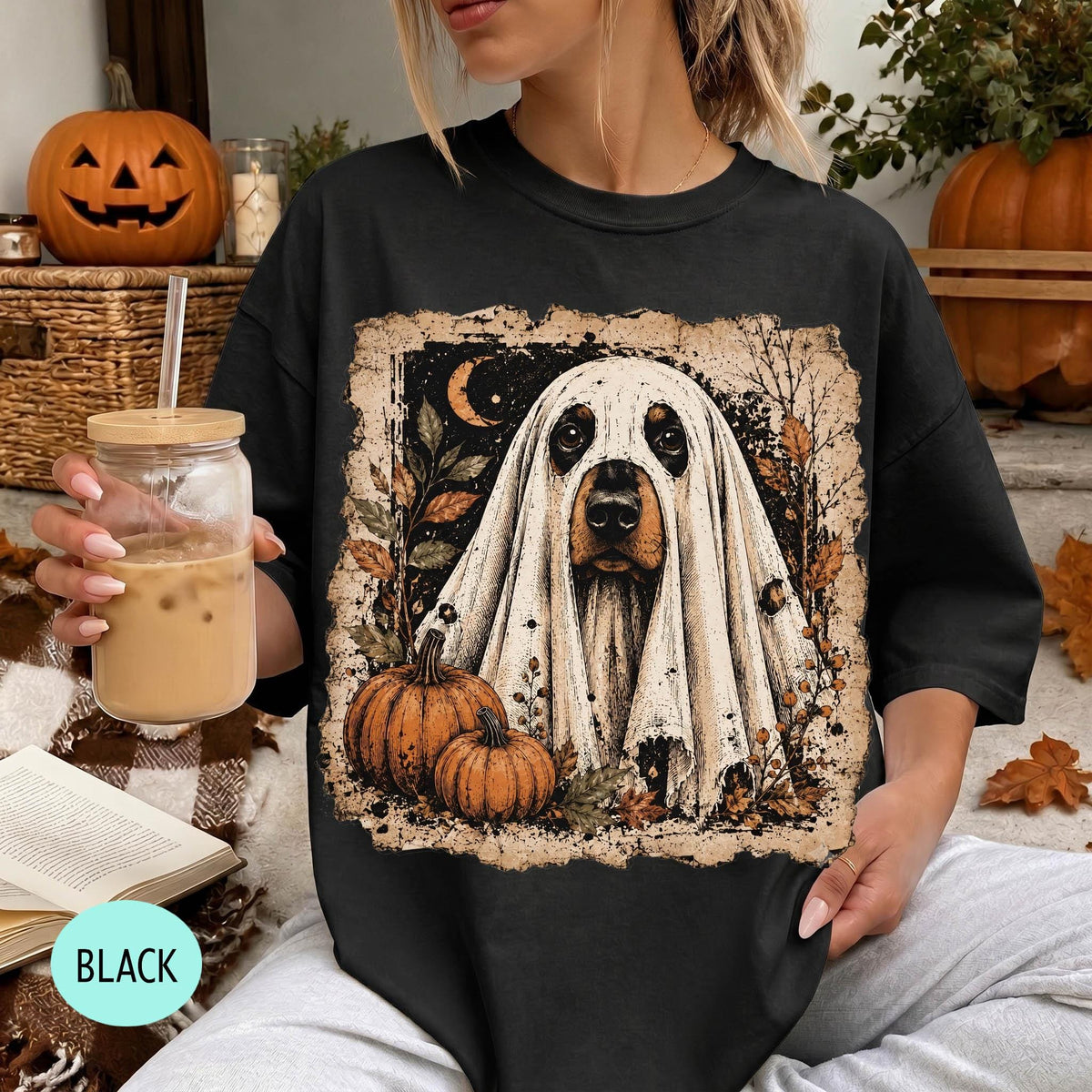 A woman wearing a black t-shirt with a Halloween-themed graphic of a ghost dog, holding a drink and surrounded by pumpkins and a jack-o-lantern.