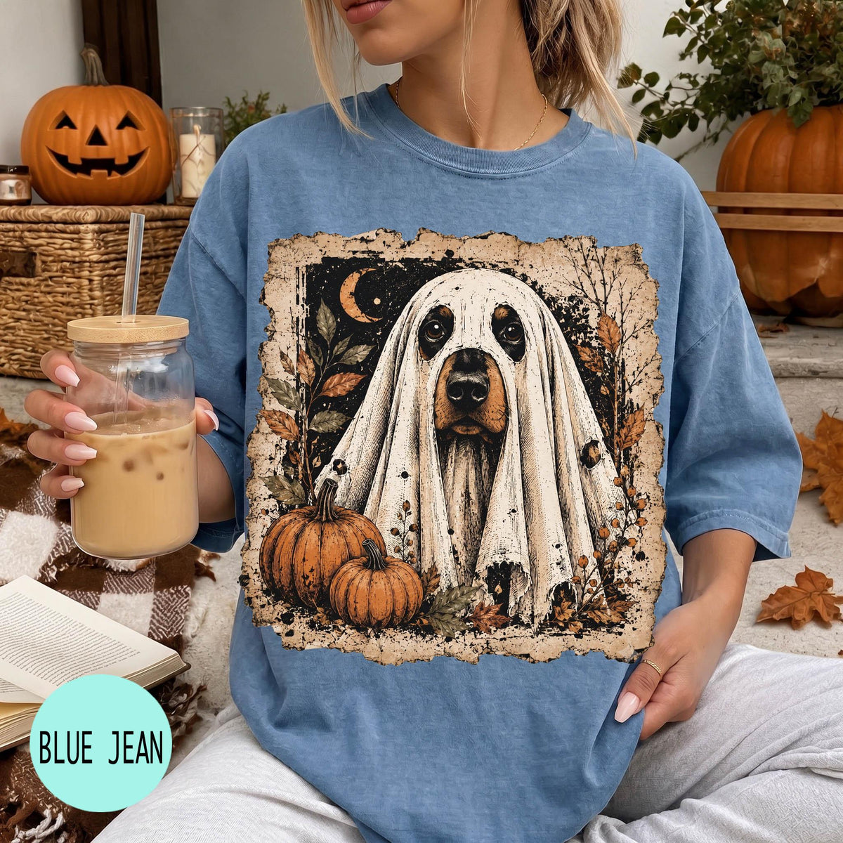 A woman is sitting on a couch, wearing a blue t-shirt with a Halloween-themed graphic of a ghost dog. She is holding a drink in a glass with a straw. There are pumpkins and a jack-o-lantern in the background, suggesting a Halloween setting.