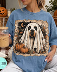 A woman is sitting on a couch, wearing a blue t-shirt with a Halloween-themed graphic of a ghost dog. She is holding a drink in a glass with a straw. There are pumpkins and a jack-o-lantern in the background, suggesting a Halloween setting.