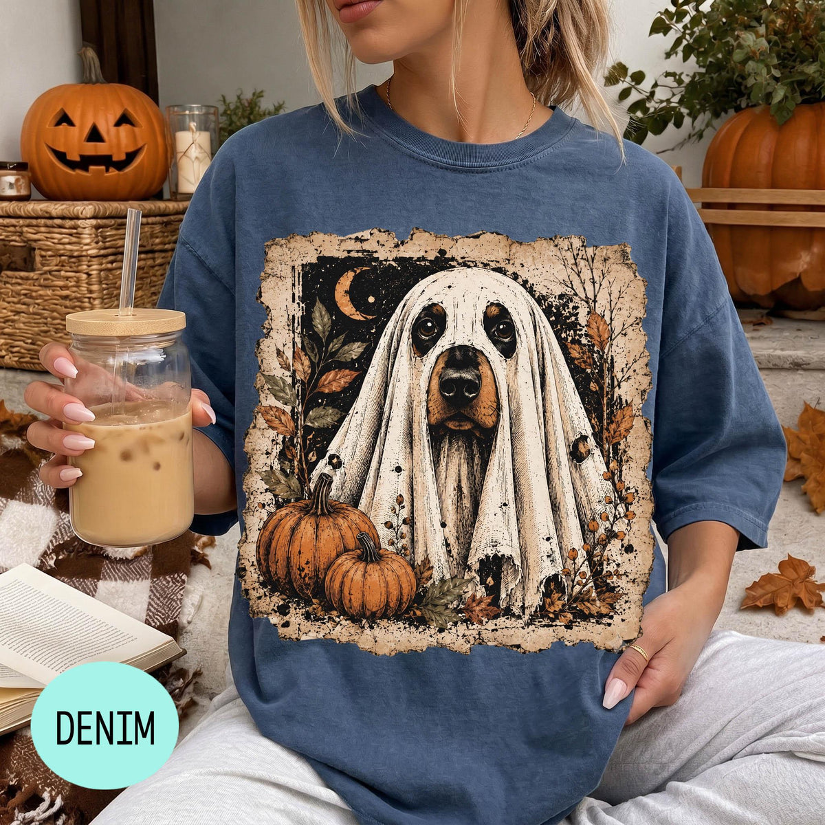 A woman wearing a blue t-shirt with a graphic of a ghost dog and pumpkins, holding a drink and surrounded by Halloween decorations.