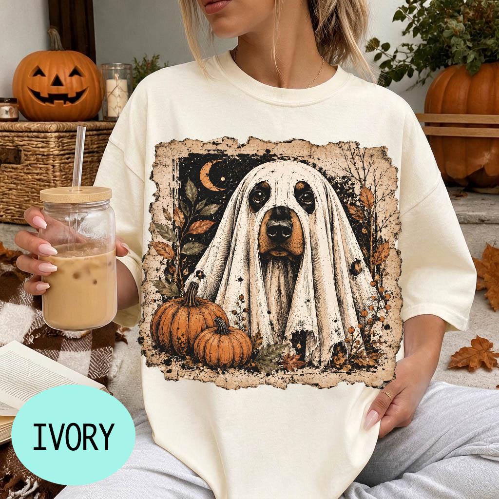 A woman wearing a white t-shirt with a graphic of a ghost dog and pumpkins, holding a glass of iced coffee.