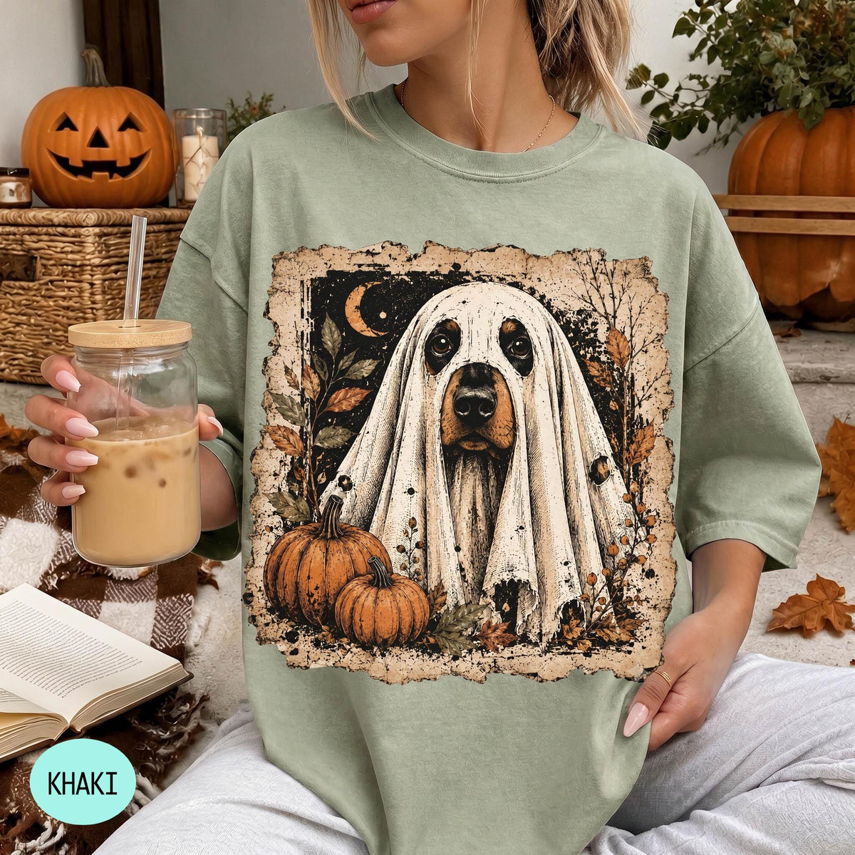 A woman is sitting on a couch, wearing a green t-shirt with a Halloween-themed graphic of a ghost dog. She is holding a glass of iced coffee and has a book and a pumpkin nearby.