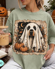 A woman is sitting on a couch, wearing a green t-shirt with a Halloween-themed graphic of a ghost dog. She is holding a glass of iced coffee and has a book and a pumpkin nearby.