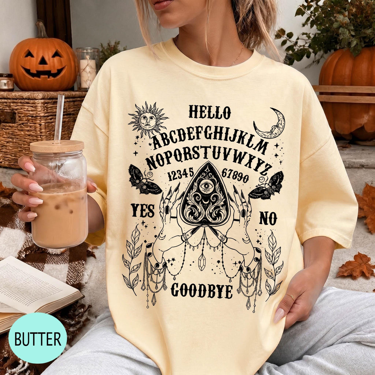A woman wearing a yellow t-shirt with a Ouija board design and text printed on it, holding a drink in a glass.