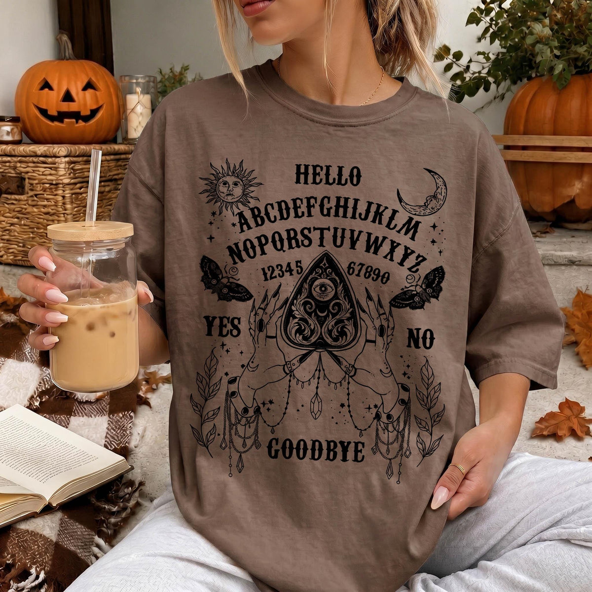 A woman wearing a t-shirt with an Ouija board design and holding a drink is sitting in a room decorated for Halloween.