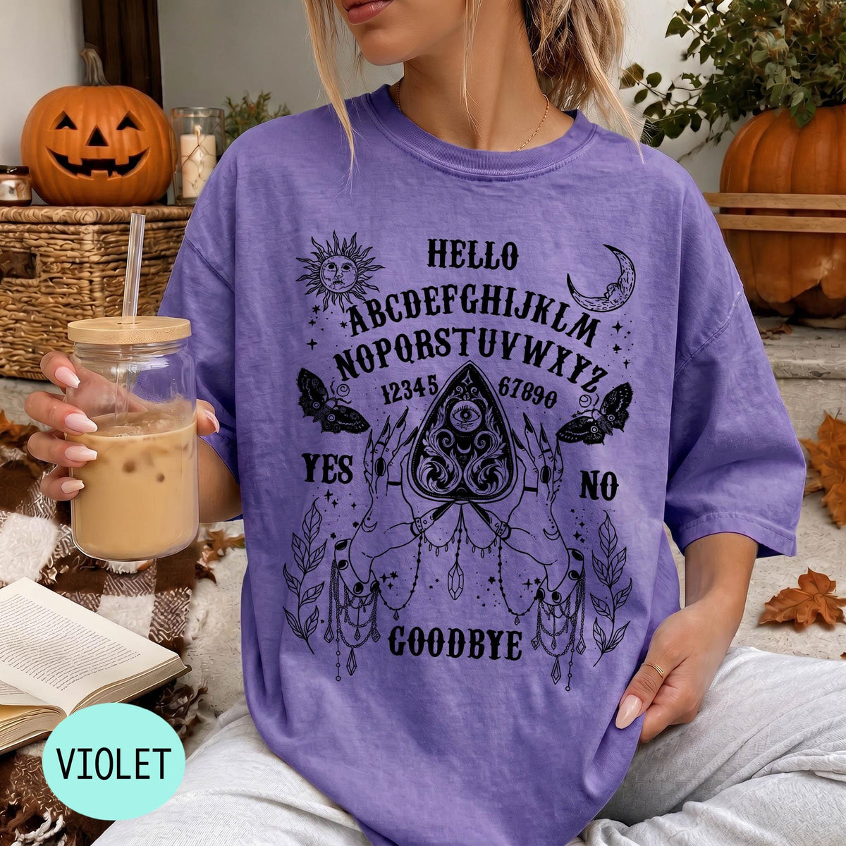 A woman wearing a purple t-shirt with an Ouija board design and holding a drink, with a pumpkin and autumn leaves in the background.