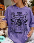 A woman wearing a purple t-shirt with an Ouija board design and holding a drink, with a pumpkin and autumn leaves in the background.