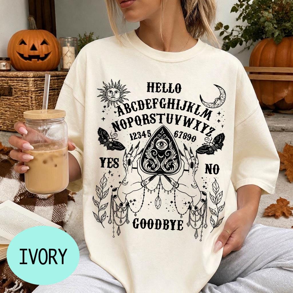 A woman wearing a white t-shirt with a Ouija board design and text printed on it, holding a drink in a glass.