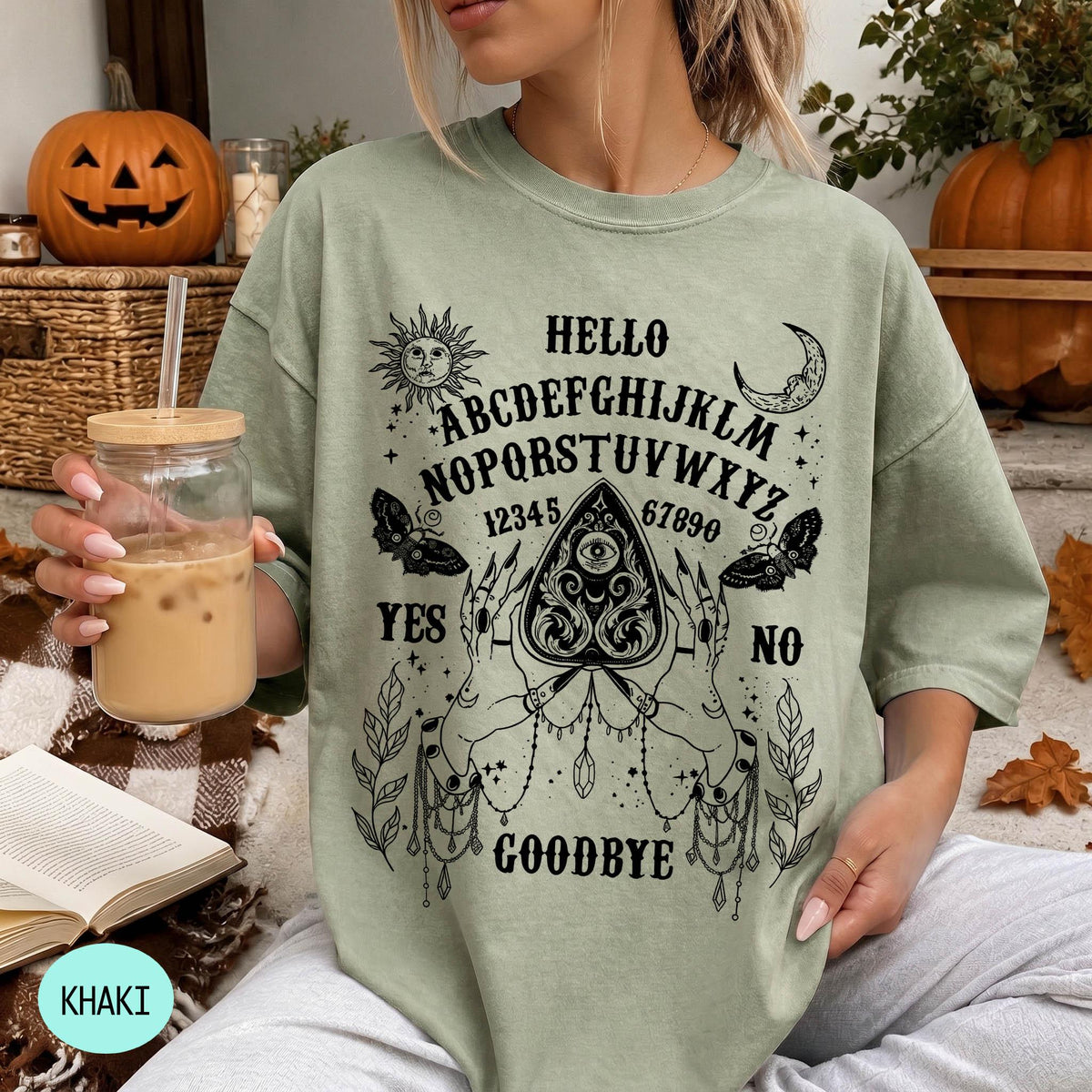 A woman wearing a green t-shirt with an Ouija board design and text printed on it, holding a drink in a glass.