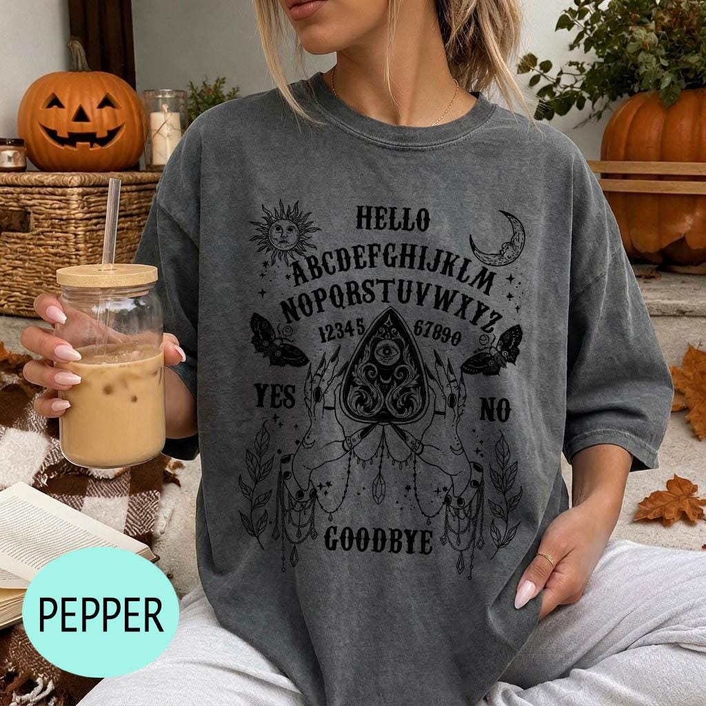 A woman wearing a gray t-shirt with a graphic design of an Ouija board and text printed on it, holding a drink in a glass.