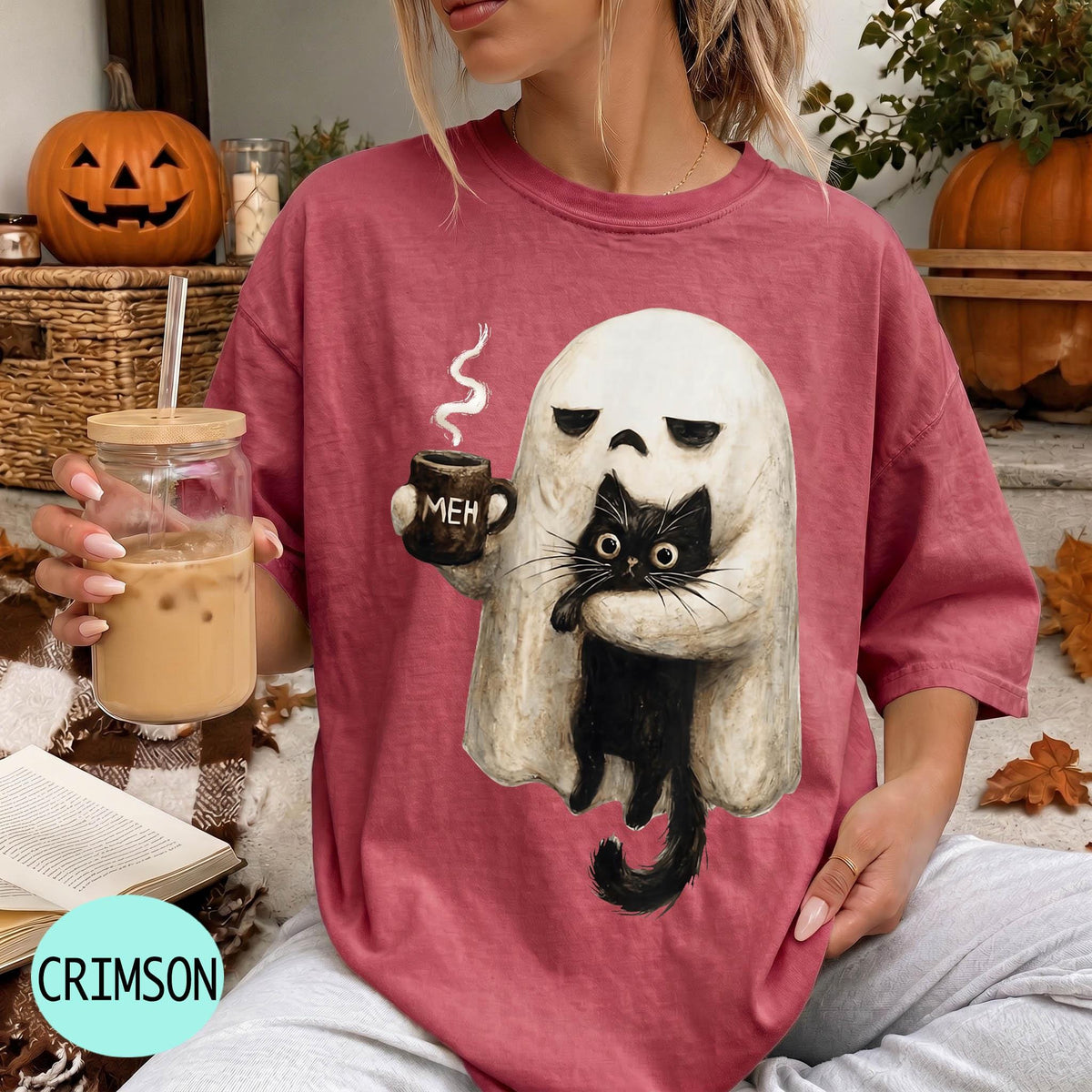 A woman wearing a red t-shirt with a graphic of a ghost holding a coffee mug and a black cat, sitting in front of a pumpkin and a book.