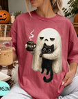 A woman wearing a red t-shirt with a graphic of a ghost holding a coffee mug and a black cat, sitting in front of a pumpkin and a book.