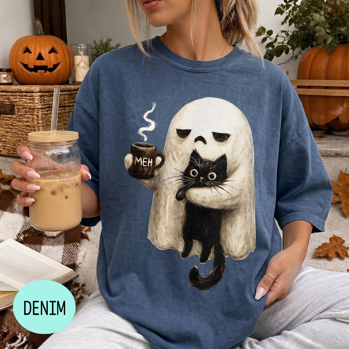 A woman wearing a blue t-shirt with a graphic of a ghost holding a coffee mug and a black cat, sitting on a couch with a pumpkin and a book nearby.