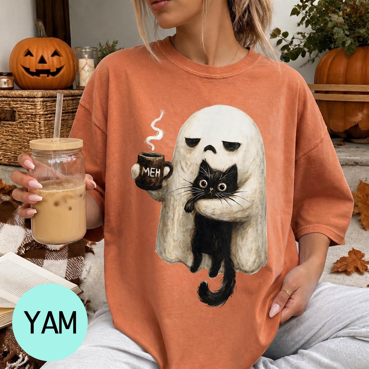 A woman wearing a t-shirt with a graphic of a ghost holding a coffee mug and a black cat, sitting on a couch with a pumpkin and a book nearby.
