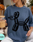 A woman wearing a blue t-shirt with a skeleton dog graphic is sitting on a couch, holding a drink in a glass with a straw.