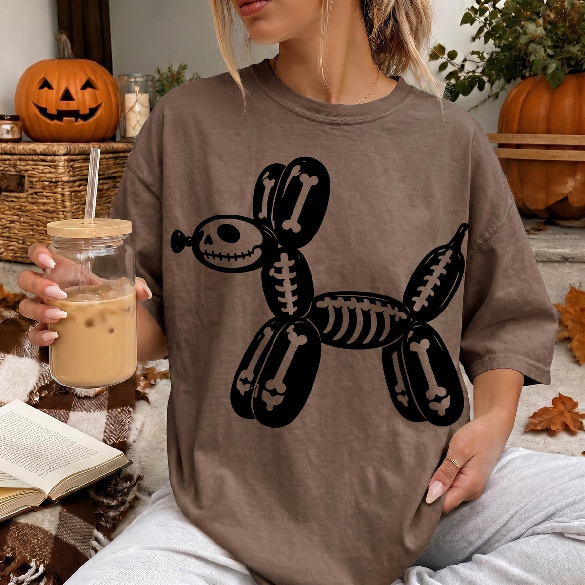 A woman is sitting on a blanket, wearing a t-shirt with a skeleton dog design, and holding a glass of iced coffee.