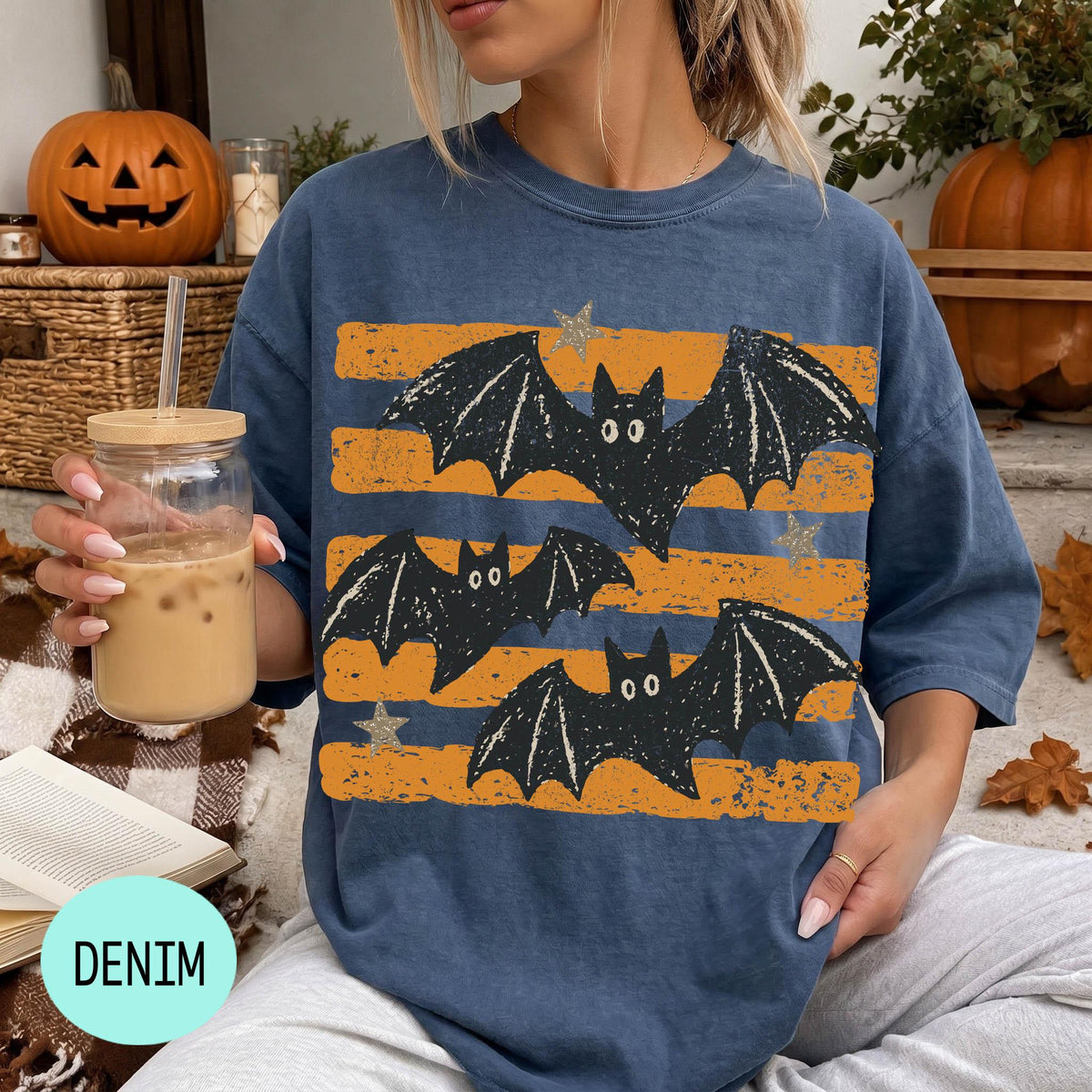 A woman wearing a blue t-shirt with a bat design is sitting on a couch, holding a drink in a glass with a straw. There are pumpkins and a book nearby, suggesting a cozy, autumnal setting.