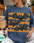 A woman wearing a blue t-shirt with a bat design is sitting on a couch, holding a drink in a glass with a straw. There are pumpkins and a book nearby, suggesting a cozy, autumnal setting.