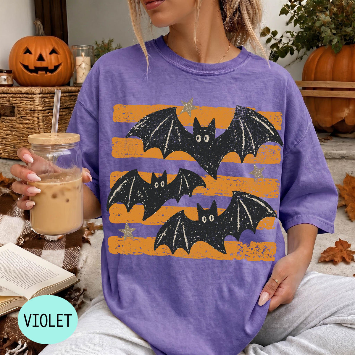 A woman wearing a purple t-shirt with a bat design is sitting on a couch, holding a drink in a glass. There are pumpkins and a book visible in the background.