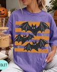 A woman wearing a purple t-shirt with a bat design is sitting on a couch, holding a drink in a glass. There are pumpkins and a book visible in the background.