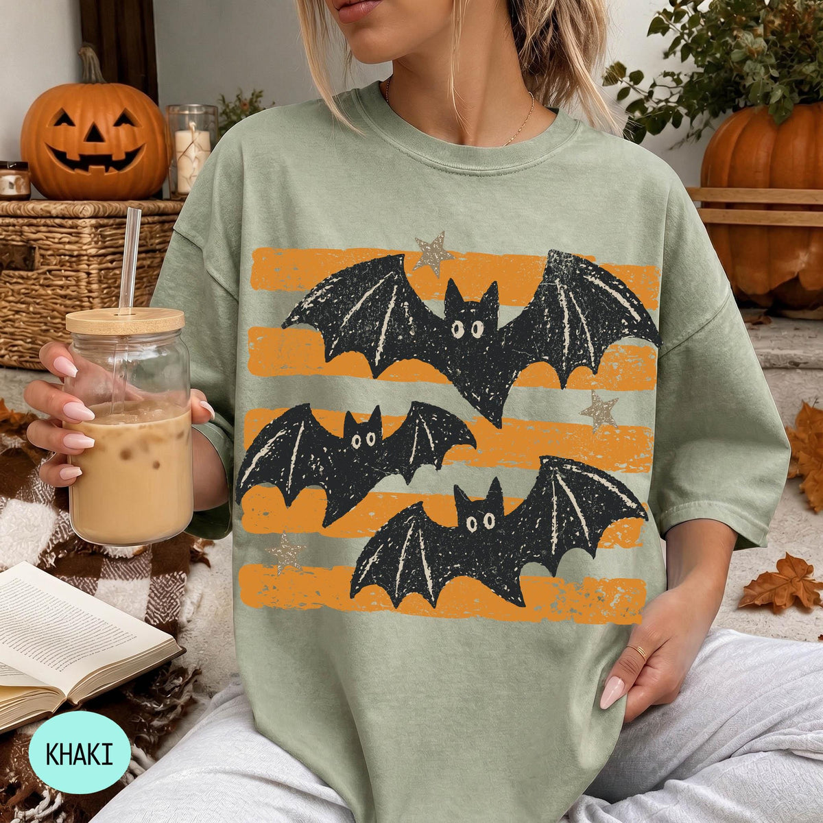 A woman is sitting on a couch, wearing a green t-shirt with a graphic of bats and stars. She is holding a glass of iced coffee.