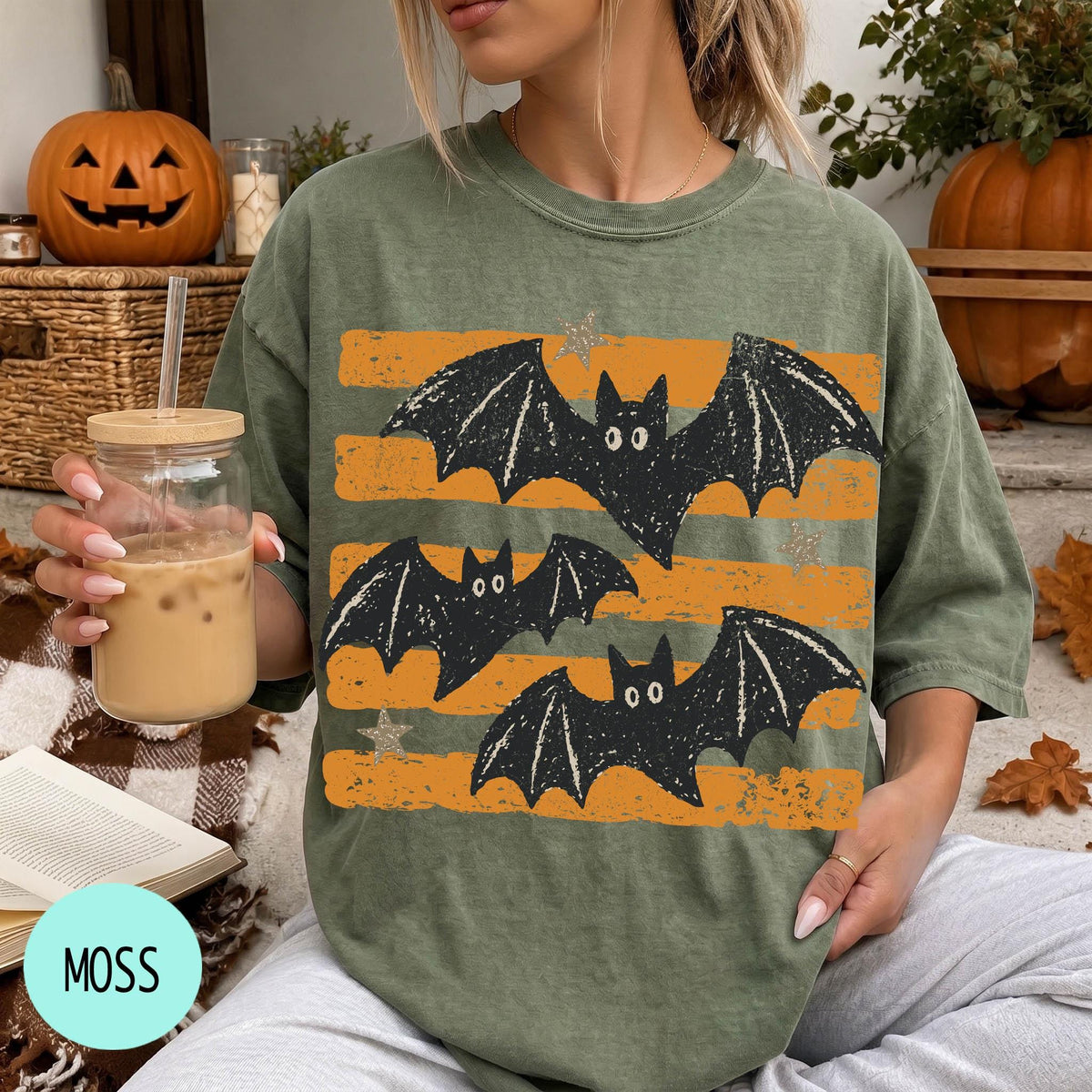 A woman is sitting on a couch, wearing a green t-shirt with a graphic of bats and orange stripes. She is holding a drink in a glass with a straw.