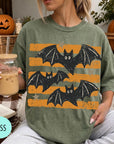 A woman is sitting on a couch, wearing a green t-shirt with a graphic of bats and orange stripes. She is holding a drink in a glass with a straw.